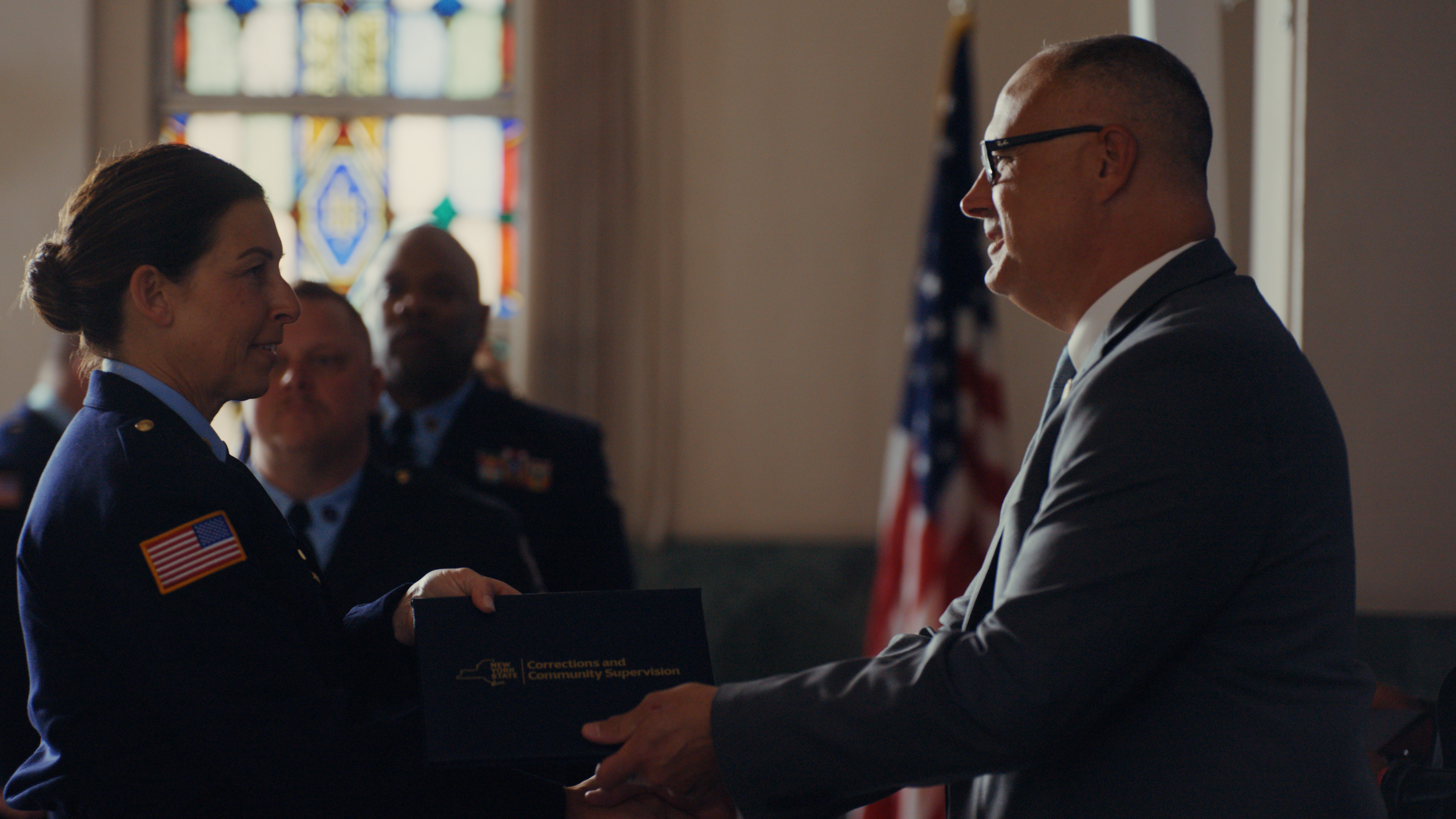 The commissioner of the New York Department of Corrections presents a certificate to a female officer with the U.S. flag and stained glass behind them.