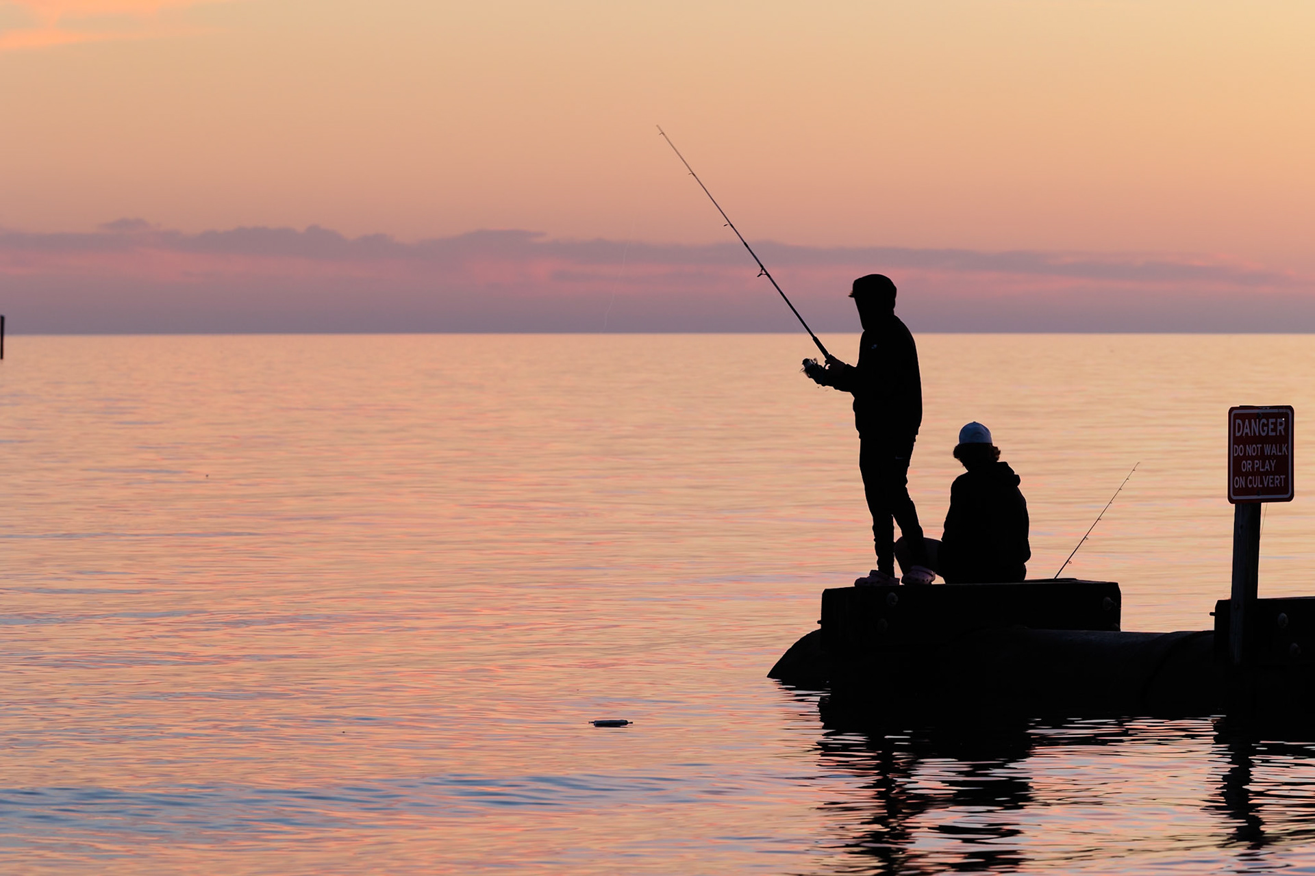 220114_246 Silhouette of young adults ignoring danger sign while fishing on storm drain culvert in Long Beach, Mississippi, USA