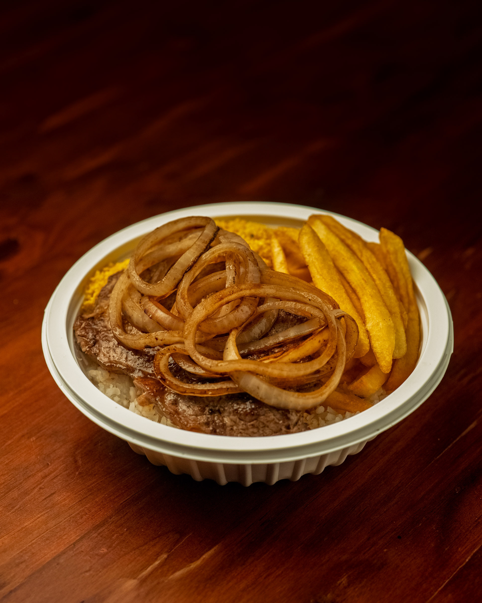 Marmita de bife acebolado com farofa e batata frita, servida em ângulo que mostra o volume da embalagem.