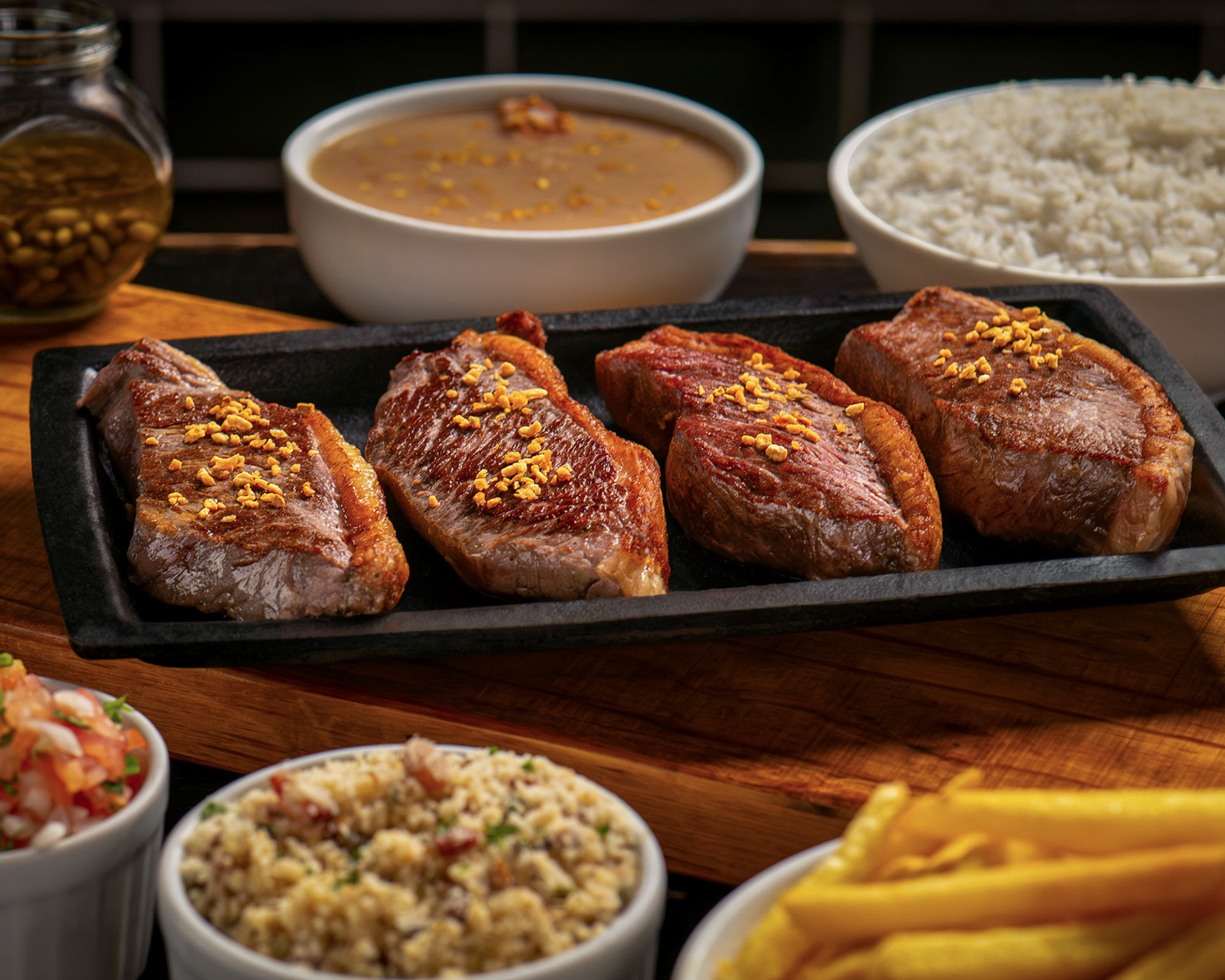 Quatro cortes de picanha grelhada com alho frito servida em travessa preta, acompanhada de arroz, feijão, farofa e vinagrete.