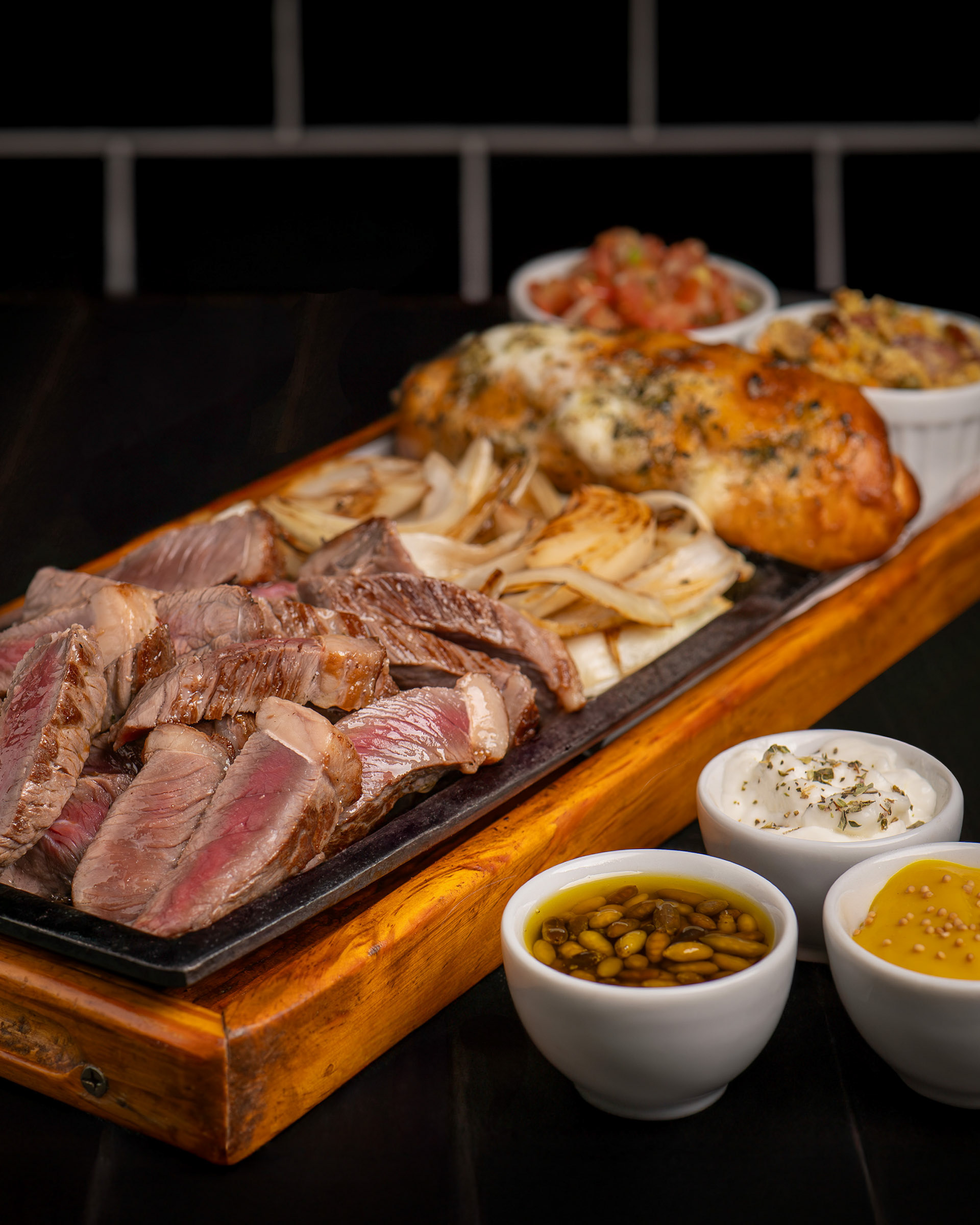 Porção de picanha fatiada e cebola grelhada servida na chapa, acompanhada de pão de alho e diversos molhos em travessa de madeira.