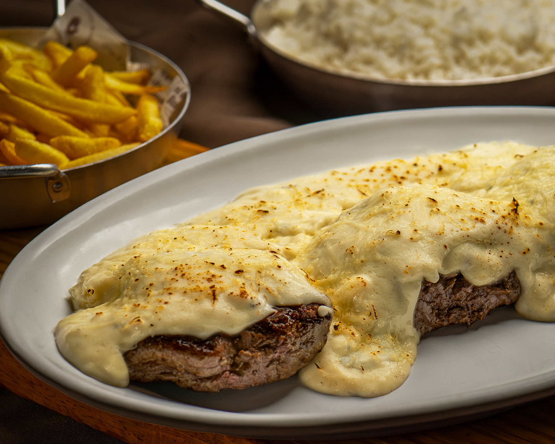 Filé mignon gratinado com queijo cremoso servido em prato branco oval, com acompanhamento de batata frita e arroz.