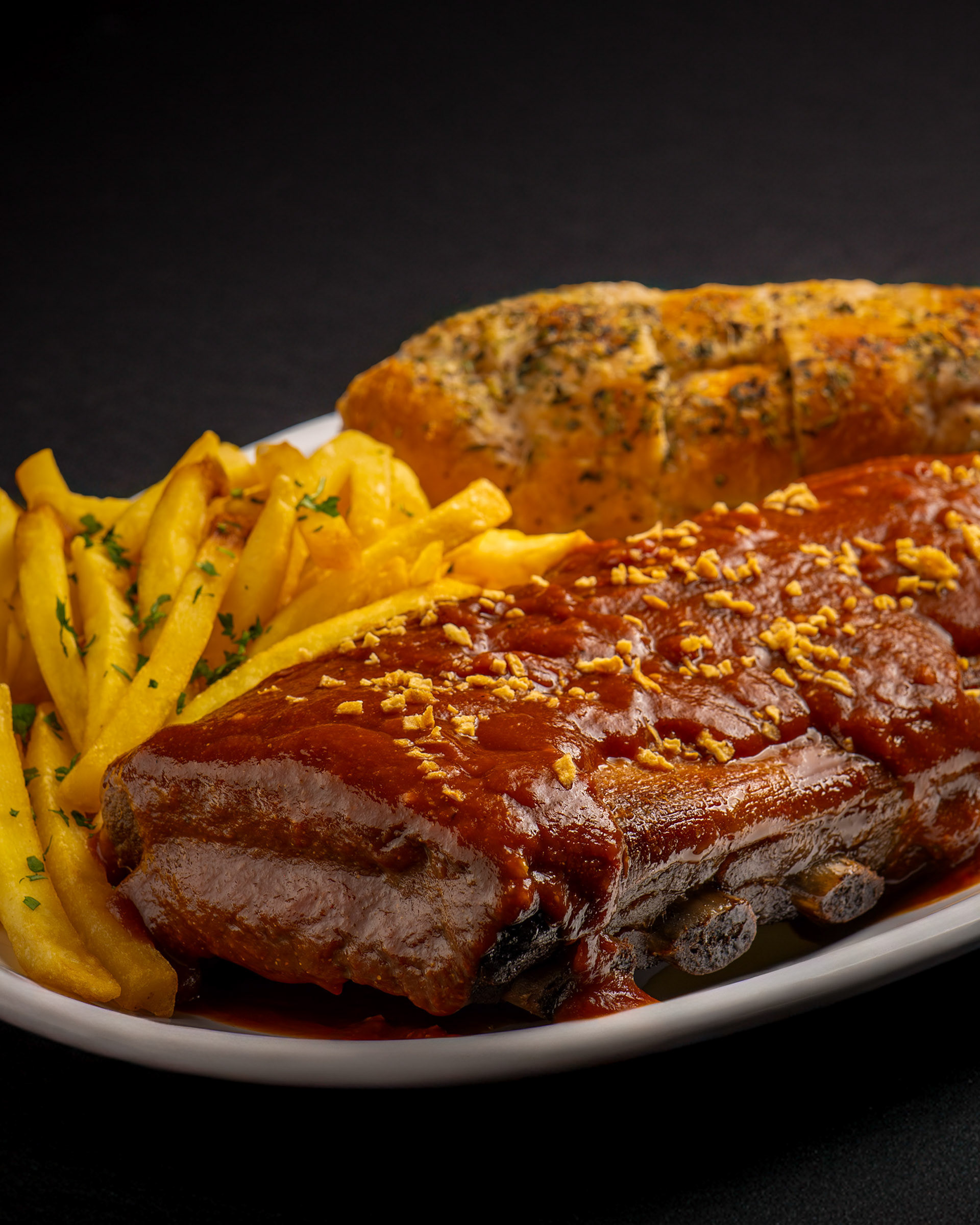 Costelinha suína com molho barbecue e alho frito, servida com batata frita com salsinha e pão de alho.