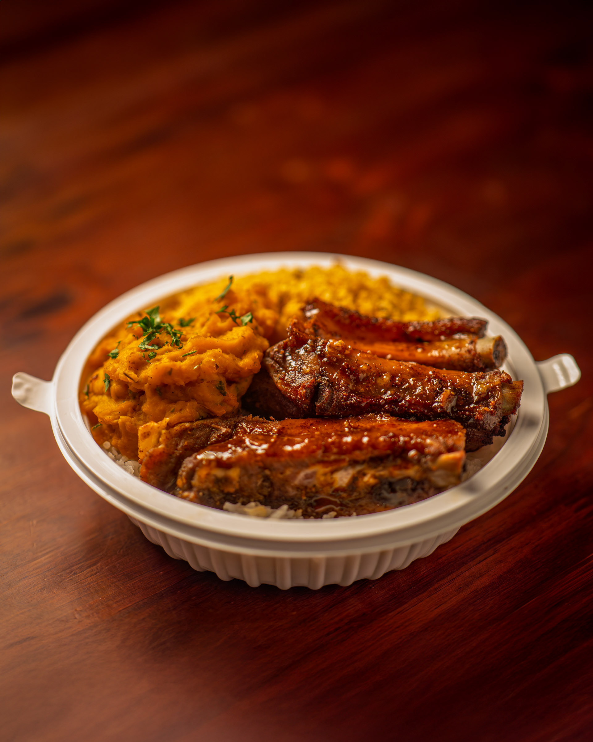 Marmita de costelinha de porco em molho barbecue brilhante, servida com purê de abóbora e arroz branco em embalagem para viagem.