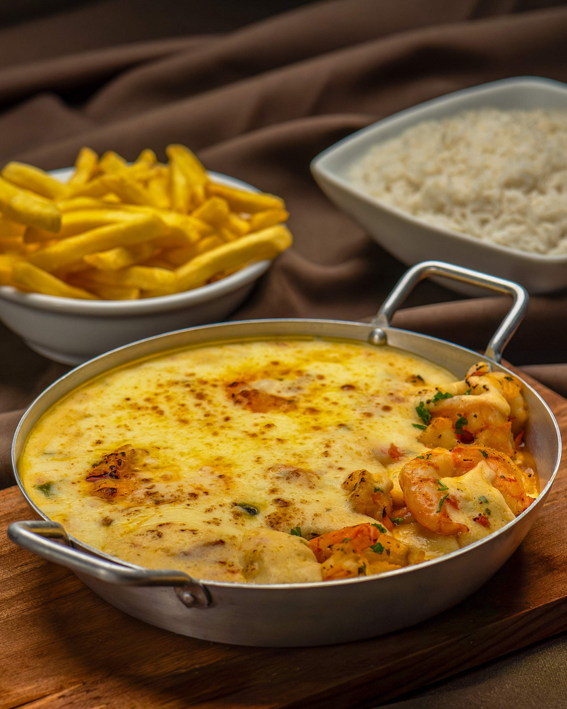 Camarão gratinado com queijo cremoso em travessa de metal, acompanhado por arroz branco e batata frita.