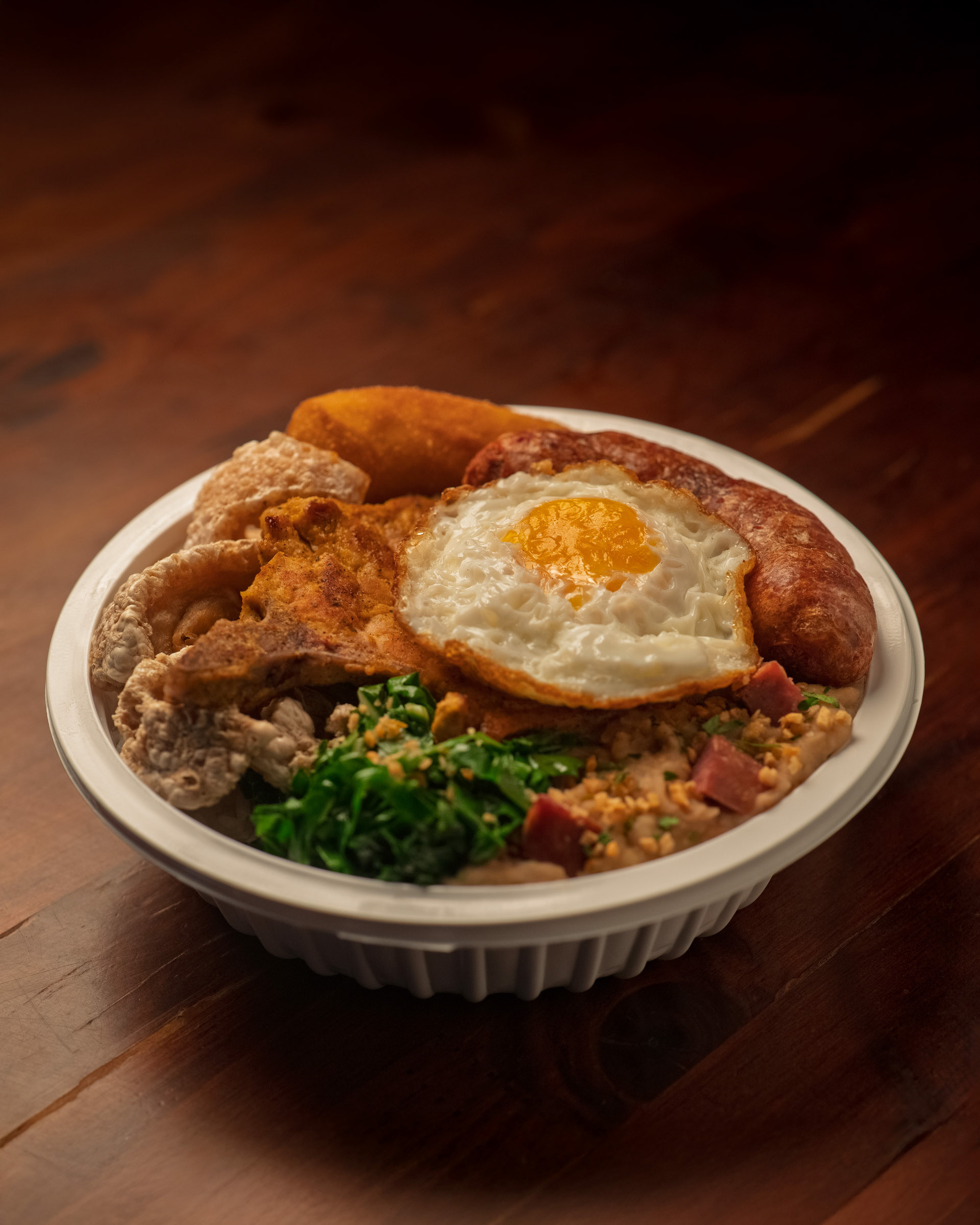 Marmita de Virado à Paulista com ovo frito, linguiça, torresmo, couve e carne, servido em embalagem de isopor sobre madeira.