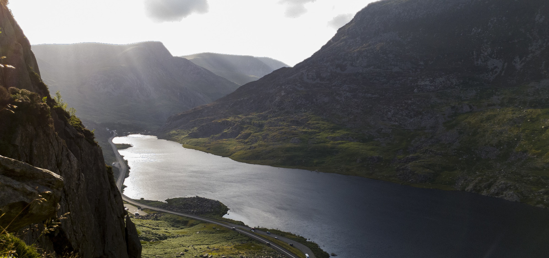 Sunlight breaking through clouds above a mountain lake and winding road.
