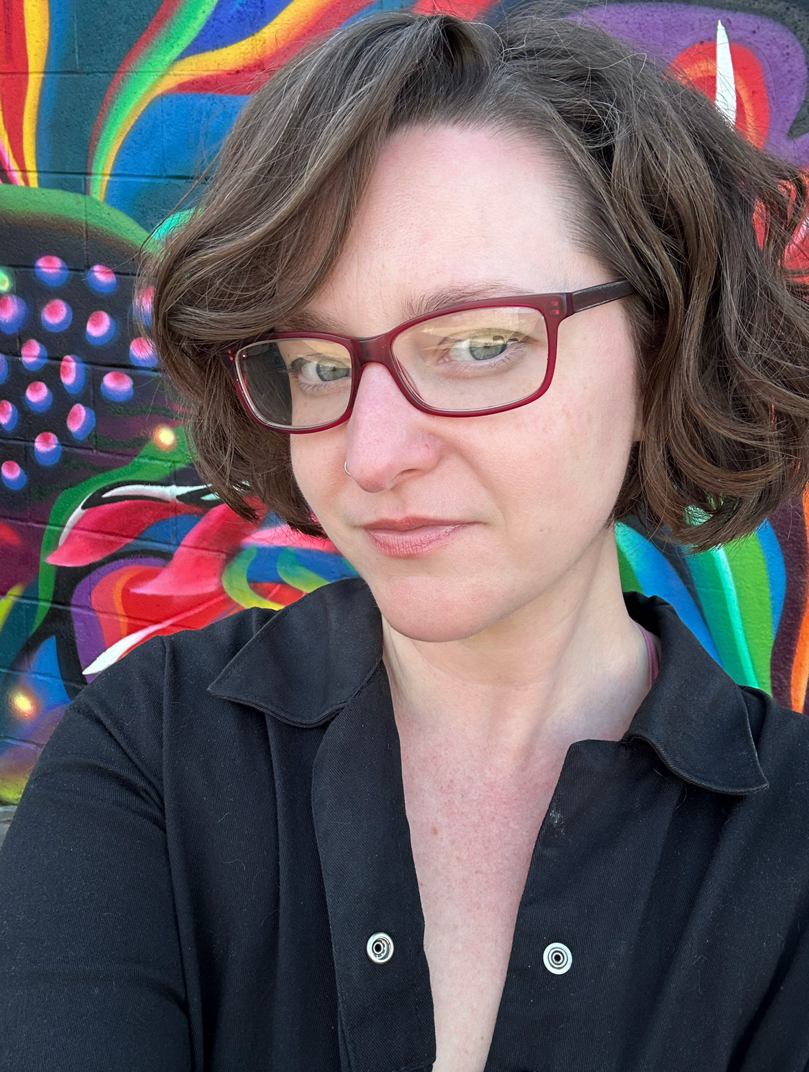 Lauren wearing glasses and black coveralls standing in front of colorful abstract botanical mural
