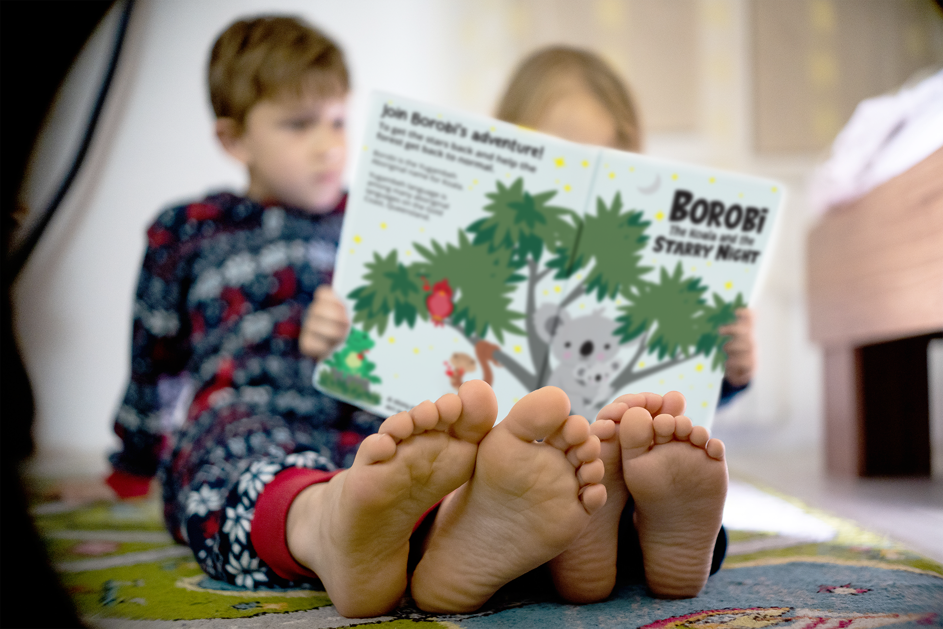 Two children sit together on the floor, focused on reading the children's book 'Borobi the Koala and the Starry Night'. The image captures their bare feet in the foreground, with the book held up close to their faces. The book's cover features a koala in a tree under a starry sky, creating a cosy and engaging reading moment.