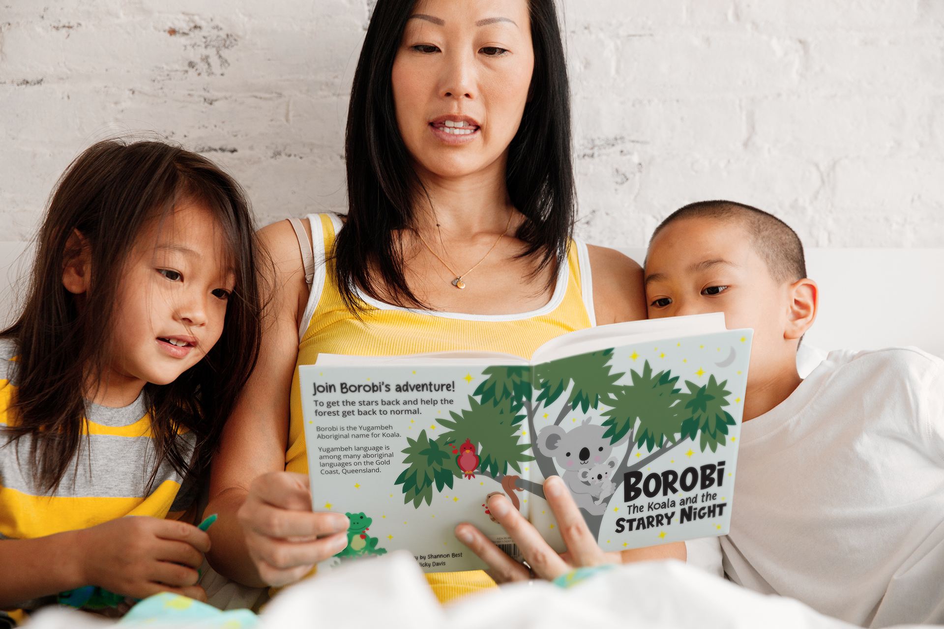 A mother reads the children's book 'Borobi the Koala and the Starry Night' to her two young children, who are closely engaged with the story. The cover of the book features a koala in a eucalyptus tree under a starry sky. The family is seated together, creating a warm and intimate reading moment, with the mother and children dressed in casual, comfortable clothing.