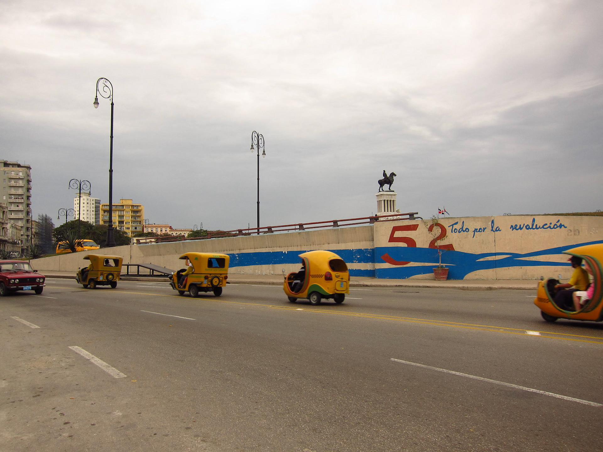 Tuk Tuk taxis zoom by a sign commemorating Cuba's communist revolution