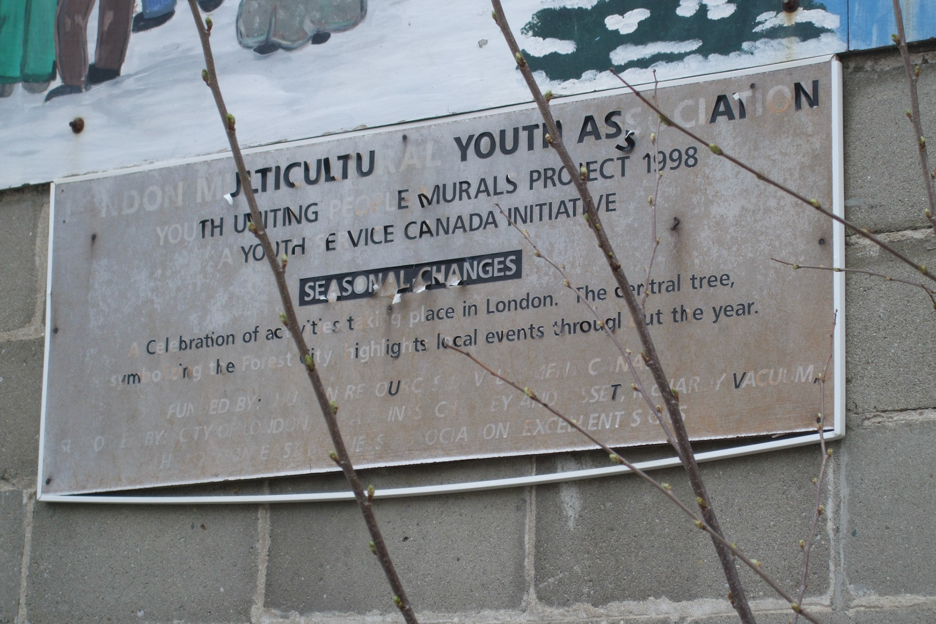 Seasonal Changes indeed... a neglected sign marking a wall mural, with budding tree branches in front.