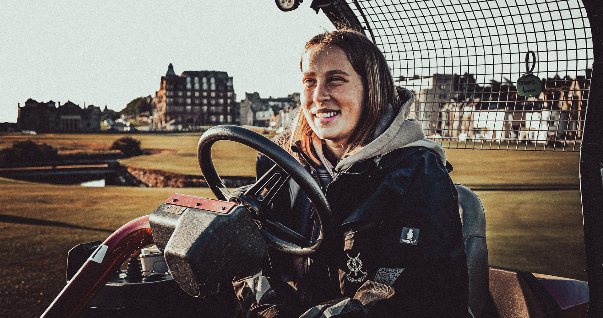 Young apprentice greenkeeper sits on top of lawnmower at dawn answering interview questions in a commercial sports film about golf in St Andrews, Scotland