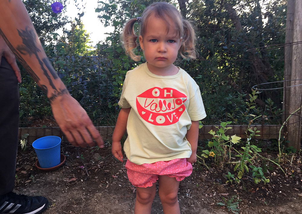 child in the garden looking glumly at camera and wearing a small Oh Vallejo Love tee.