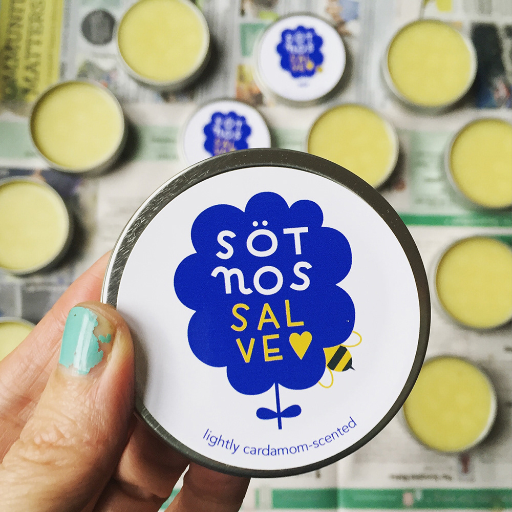 Hand holding tin close up with salve tins in the background. The lid says Sötnos Salve, lightly cardamom-scented, and has an illustrated flower with a bee entering the flower. 