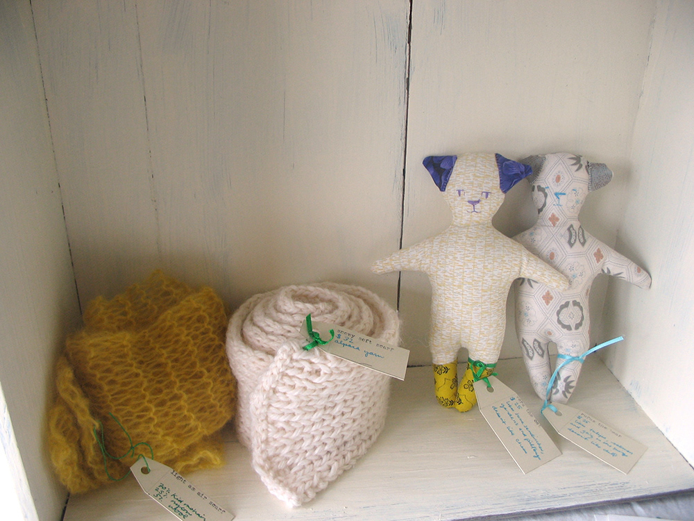A sewn cat and bear inside a cupboard next to some handknitted scarves