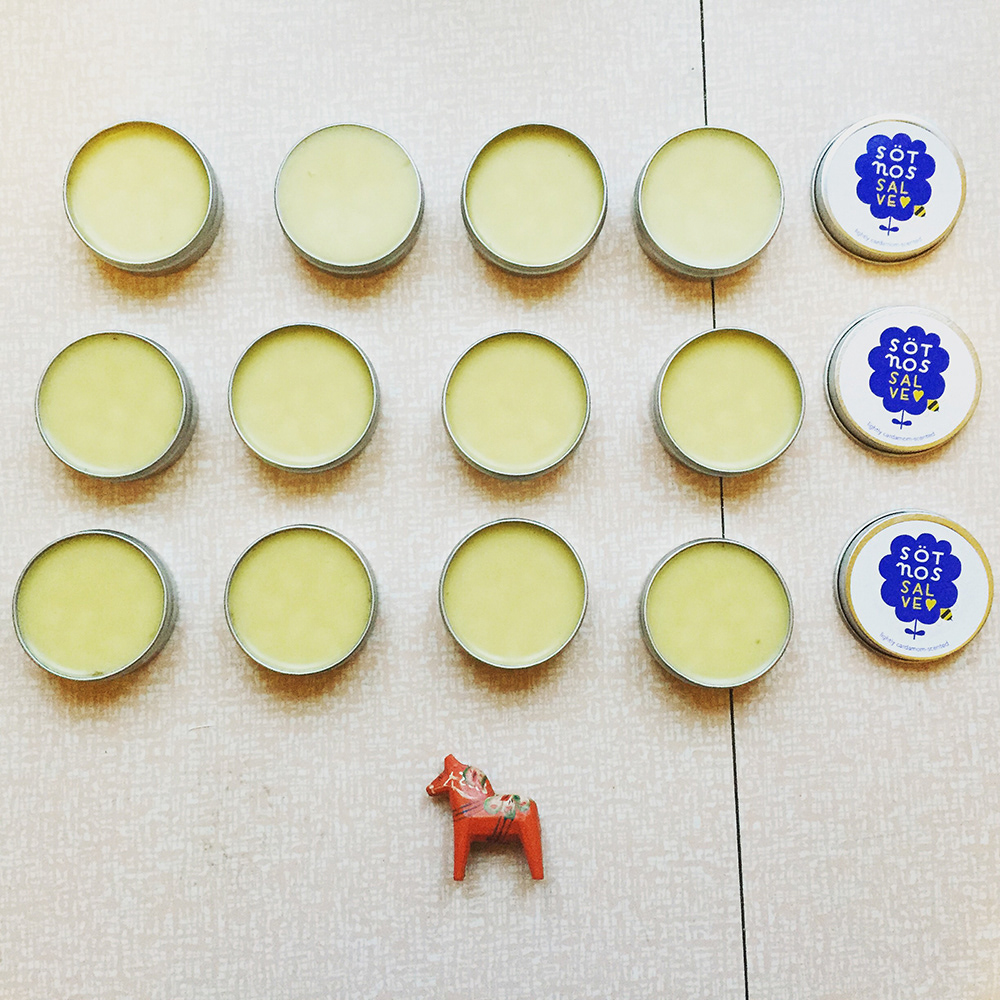 12 salve tins and 3 tin lids lined up in a grid. The tin lids say Sötnos Salve. A small Dalarna horse figurine is below the grid.
