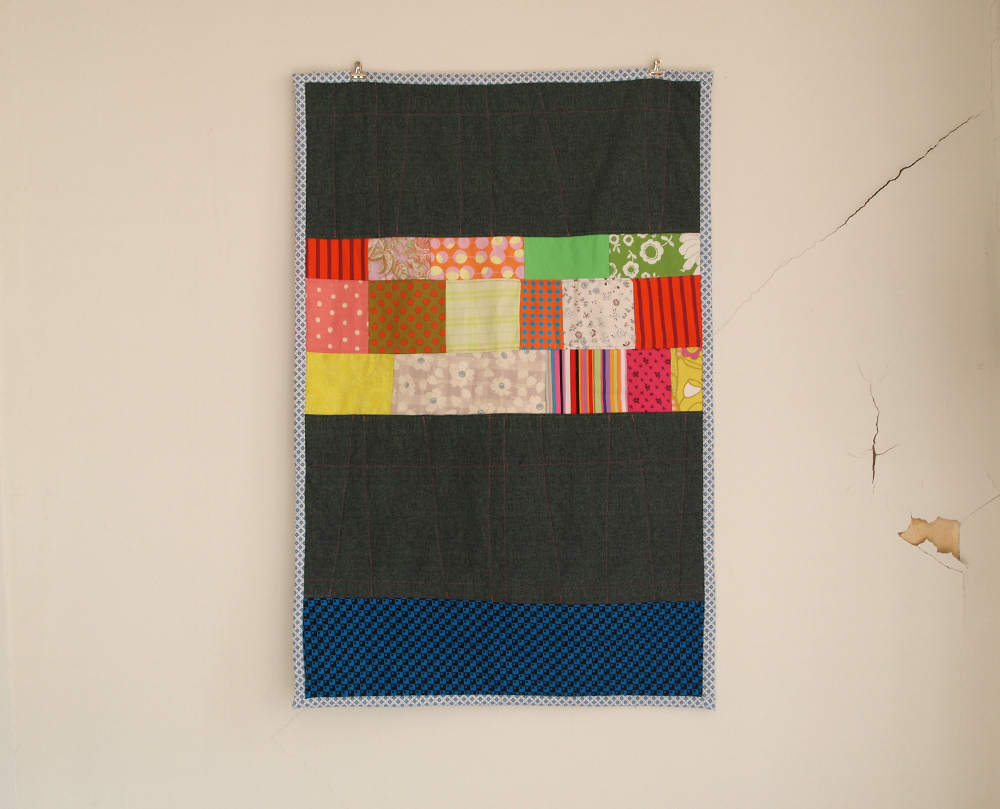 Strip quilt for a baby. It is mostly dark with three bright rows of patches in the center and a blue strip at the bottom.