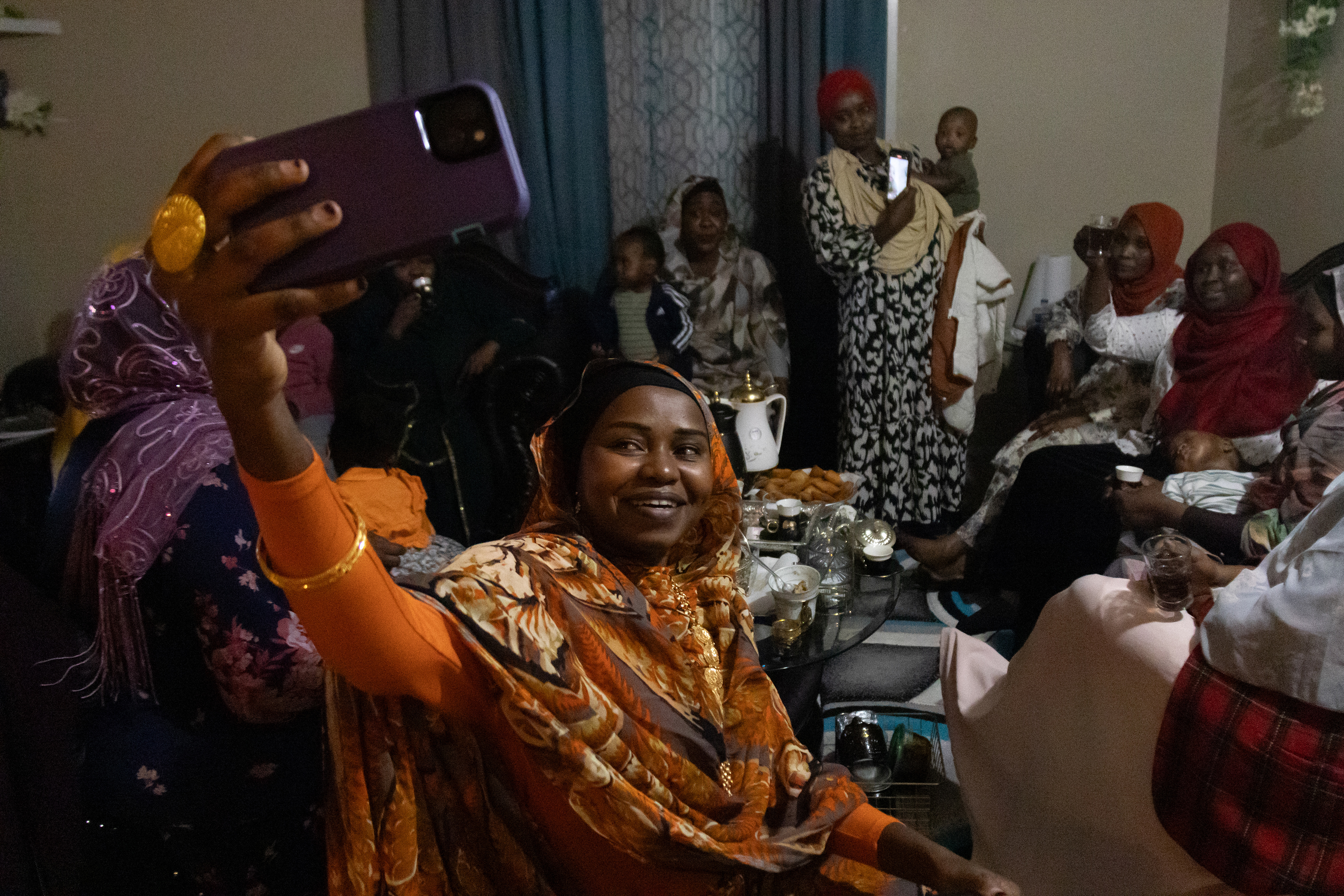 Aisha Ibrahim, holds up her phone to take a selfie with the sister get-together group in her apartment on Saturday, Feb. 23, 2025, in Lincoln, Neb. The women have found a sense of community in the group, which was formed after many of them met while visiting a friend after she had a baby in 2021. In Sudanese culture, 40 days after a woman has given birth the women of the community will visit to celebrate. 