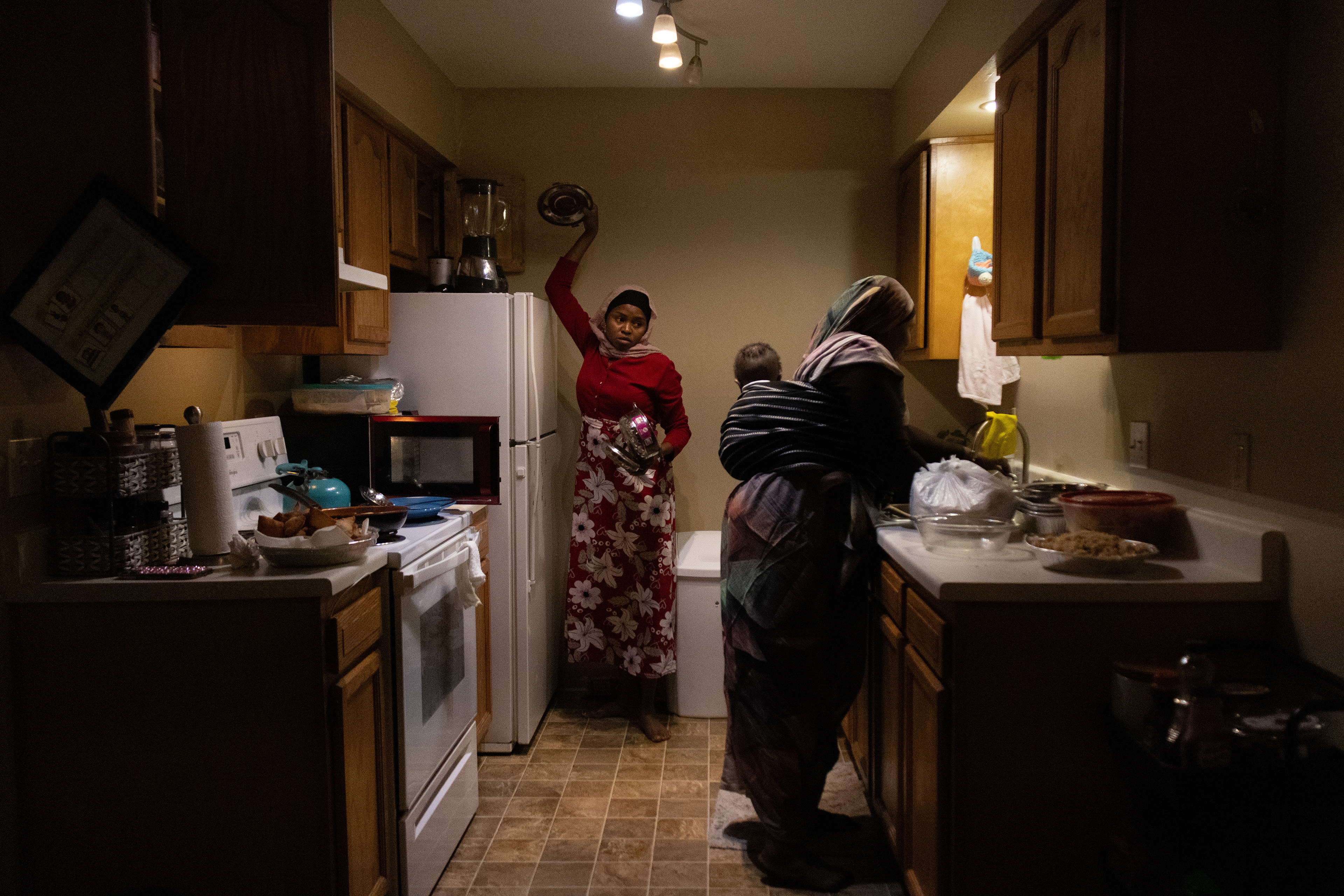 Aisha Ibrahim, left, pulls a pan lid from the top of the fridge as she talks to her sister-in-law, Halima Fadul, right, who has her 8-month-old son, Adam Abdul, tied to her back on Saturday, Feb. 23, 2025, in Lincoln, Neb. In Sudanese culture, women carry their babies on their backs as they work. Fadul arrives early to help Ibrahim with the last few dishes before the guests arrive. 