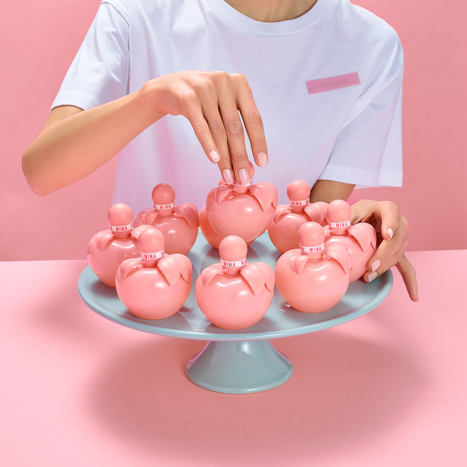 Nina Ricci Parfums : Nina Rouge, photographie de nature morte, parfum, une modèle en t-shirt Nina Ricci arrange des flacons dans une composition sur une assiette de présentation bleu ciel - PALAST PHOTOGRAPHIE, Paris France