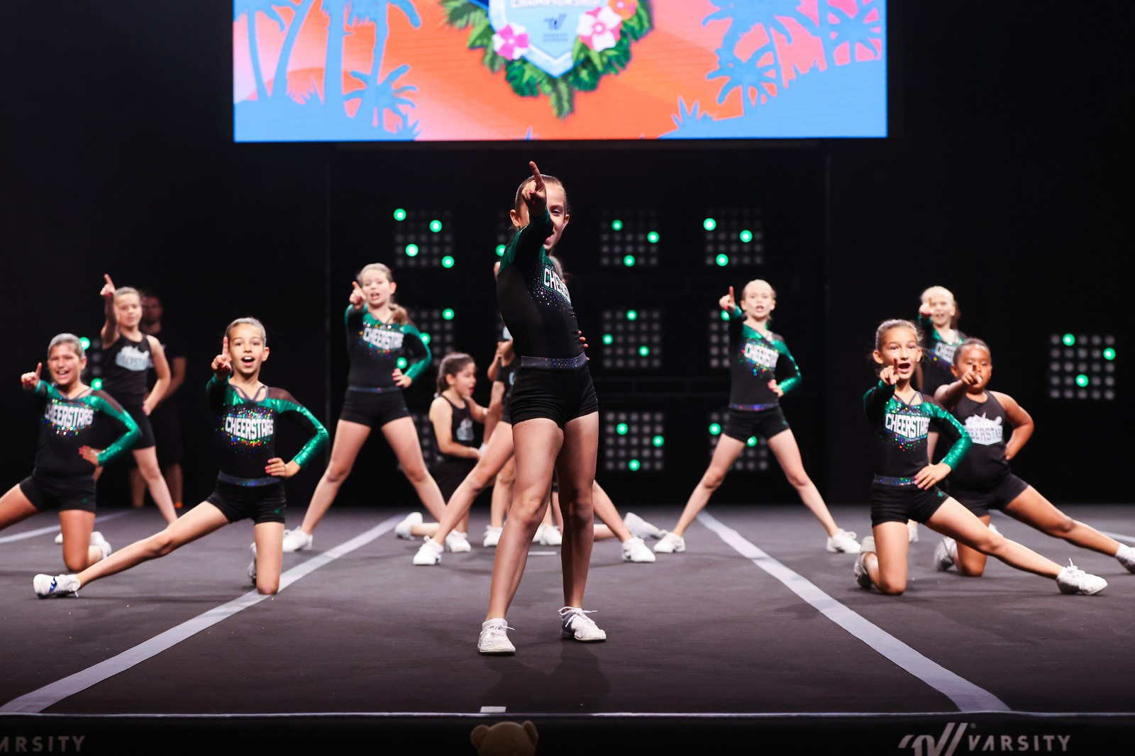 Meisterschaftsfoto von Cheerleadern aus Köln. Mädchen stehen auf Wettkampfsmatte und zeigen mit dem Finger in die Kamera.