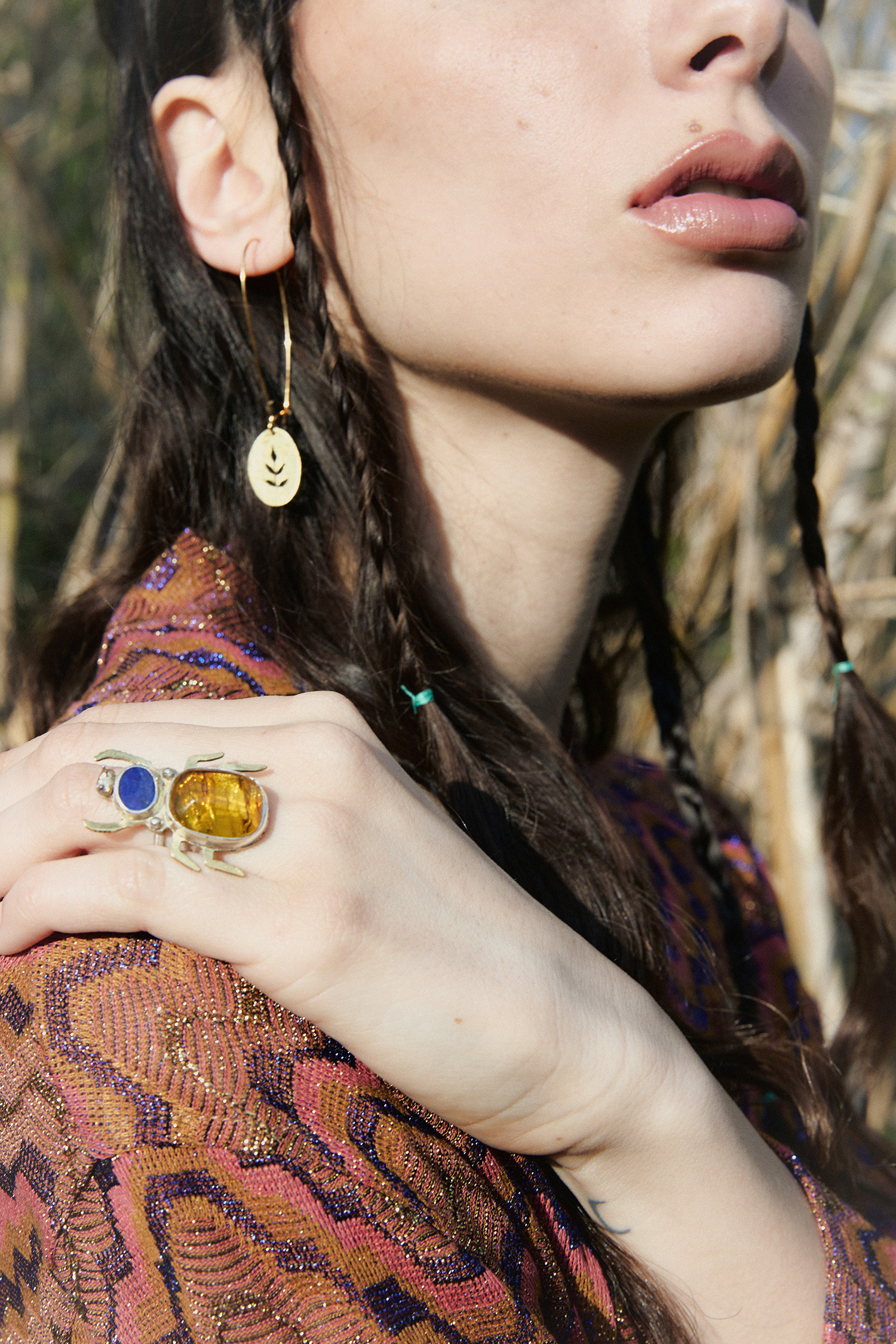 Dettaglio di volto femminile con labbra nude, treccine sottili e orecchino dorato, mano con anello scultura a forma di insetto con pietre color ambra e blu, su sfondo naturale.