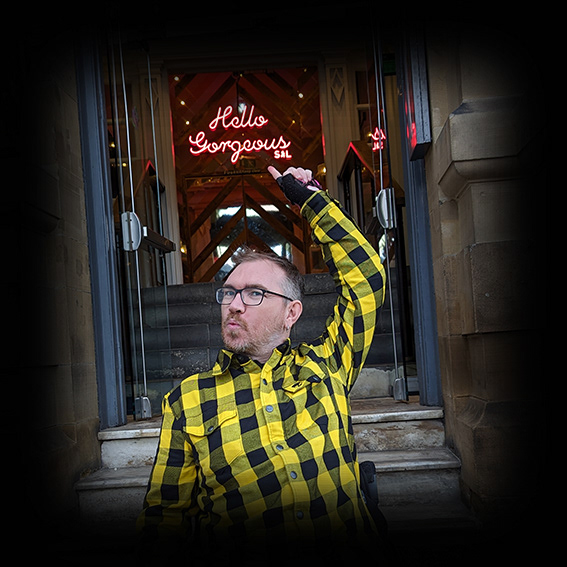 Damian, a white male wearing black framed glasses with short, mousy hair is wearing a yellow and black checked shirt. He is outside the open door of an entrance to a pub and only shown from the chest upwards. He is leaning back slightly, pointing over his left shoulder to a neon sign reading ‘Hello Gorgeous’ and his lips are puckered
