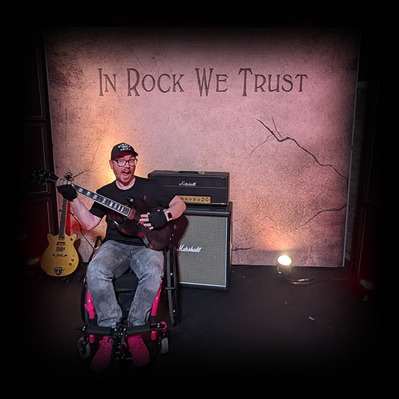Damian, a white male wearing black framed glasses with a black baseball cap in a pink framed wheelchair. He is wearing a black t-shirt, grey jeans and pink shoes. His chair is positioned in front of a photo studio backdrop made to look like stone with cracks in. There is an amplifier behind him to his left and he is holding an electric guitar. He has an excited open-mouthed gesture.