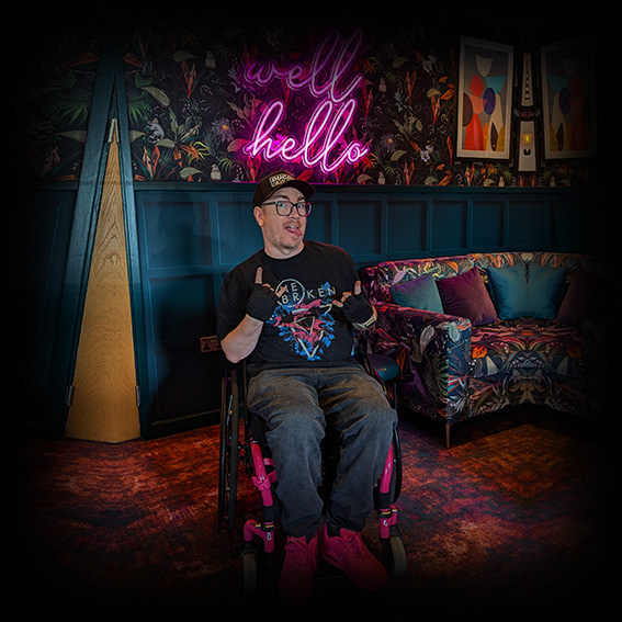 Damian, a white male wearing black framed glasses with a black baseball cap in a pink framed wheelchair. He is wearing a black tshirt, jeans and pink shoes. He is positioned in a densely patterned hotel reception area in front of a pink neon light reading ‘well hello’. He is looking at the camera, poking his tongue out of the left hand corner of his mouth and both hands are making a rock devil horns hand gesture.