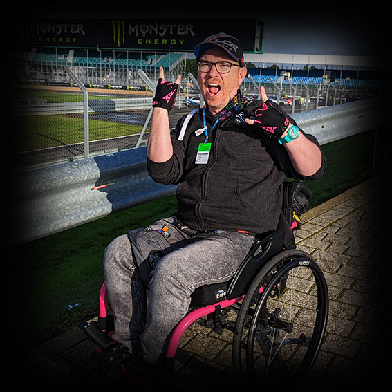 Damian, a white male wearing black framed glasses with a black LCR Honda baseball cap in a pink framed wheelchair. He is wearing a dark grey hooded top, grey jeans, black and pink fingerless gloves and pink shoes. His chair is turned slightly to the right and positioned in front of a metal crash barrier at the side of Silverstone racing circuit. His facial expression is happy and open-mouthed and both hands are making a rock devil horns hand gesture.