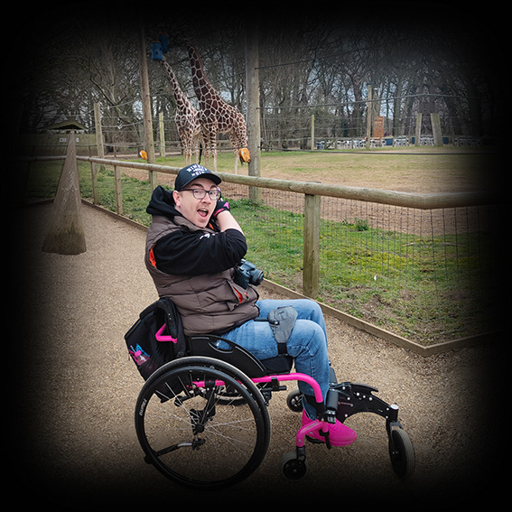 Damian, a white male wearing black framed glasses with a black baseball cap in a pink framed wheelchair. He is wearing a khaki gilet with a black hoodie underneath, blue jeans and pink shoes. He is outside at a wildlife park. He is turned with his right hand side to the camera. He is looking at the camera with a face that says ‘look!’, with both of his arms swung around to his left hand side, pointing towards 2 giraffes.