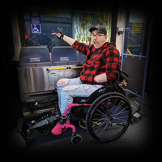 Damian, a white male wearing black framed glasses with a black baseball cap in a pink framed wheelchair. He is wearing a black and red checked hoodie, ripped blue jeans and pink shoes. His chair is positioned in a wheelchair bay on a public bus in Las Vegas, with his chair securely clamped in. The camera is to Damian’s left hand side. He has his left hand in his pocket, his right hand is pointing to a disabled logo on the bus window and he has a surprised open-mouthed look on his face, looking towards the camera