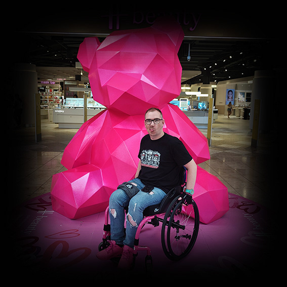 Damian, a white male wearing black framed glasses with short, mousy hair in a pink framed wheelchair. He is wearing a black tshirt, ripped blue jeans and pink shoes. He is sticking his tongue out to the camera. He is positioned in front of a giant pink geometric teddy bear sculpture in a shopping centre