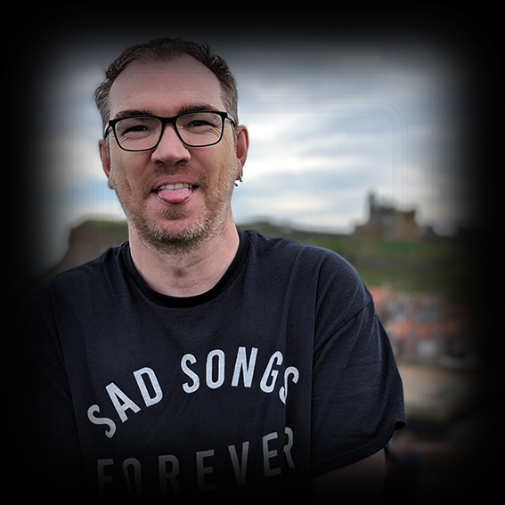 Damian, a white male wearing black framed glasses with short, mousy hair. He is wearing a black t-shirt reading Sad Songs Forever He is outside and only shown from the chest upwards. He is poking his tongue out to the camera, possibly starting to laugh
