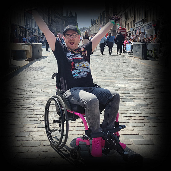 Damian, a white male wearing black framed glasses with a black baseball cap in a pink framed wheelchair. He is wearing a black tshirt, grey jeans and pink shoes. He is positioned on a cobbled street in Edinburgh in the sunshine. His chair is turned slightly to the left, his facial expression is happy and open-mouthed and he has both arms up in the air.