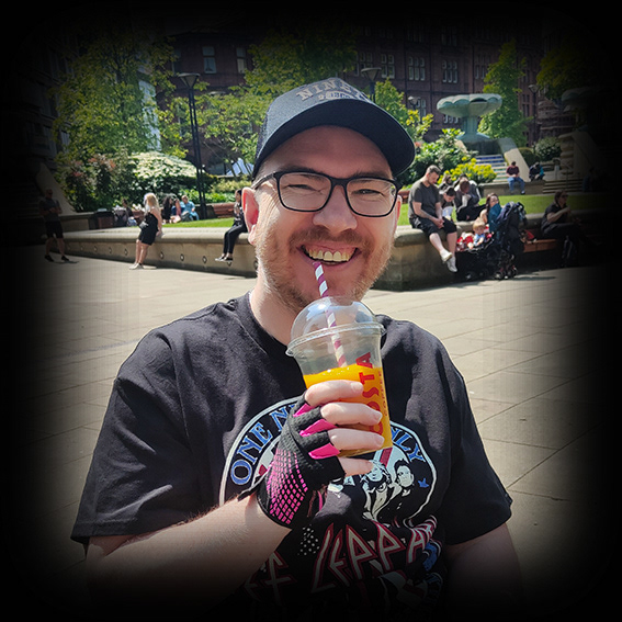 Damian, a white male wearing black framed glasses and a black baseball cap He is wearing a black Def Leppard t-shirt. His right hand is wearing a black and pink fingerless glove and holding an orange drink in a clear Costa coffee clear takeaway cup with a straw. He is outside and only shown from the chest upwards. He is smiling to the camera, possibly starting to laugh