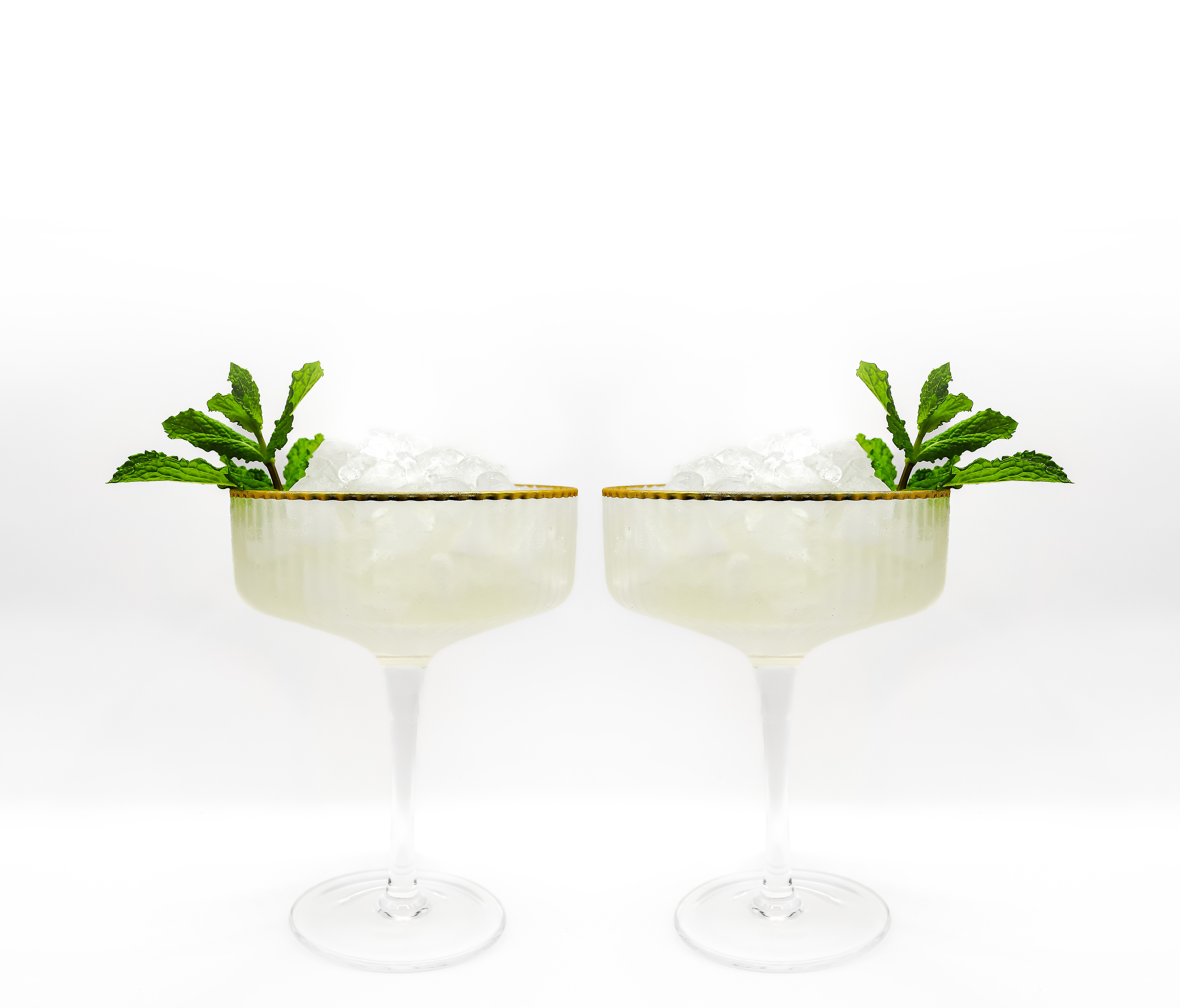 Two South Side Cocktails served in elegant coupe glasses with golden rims, filled with crushed ice and a touch of glitter visible above the glass. Each cocktail is garnished with a sprig of mint, set against a white background, making the drinks the focal point of the image.