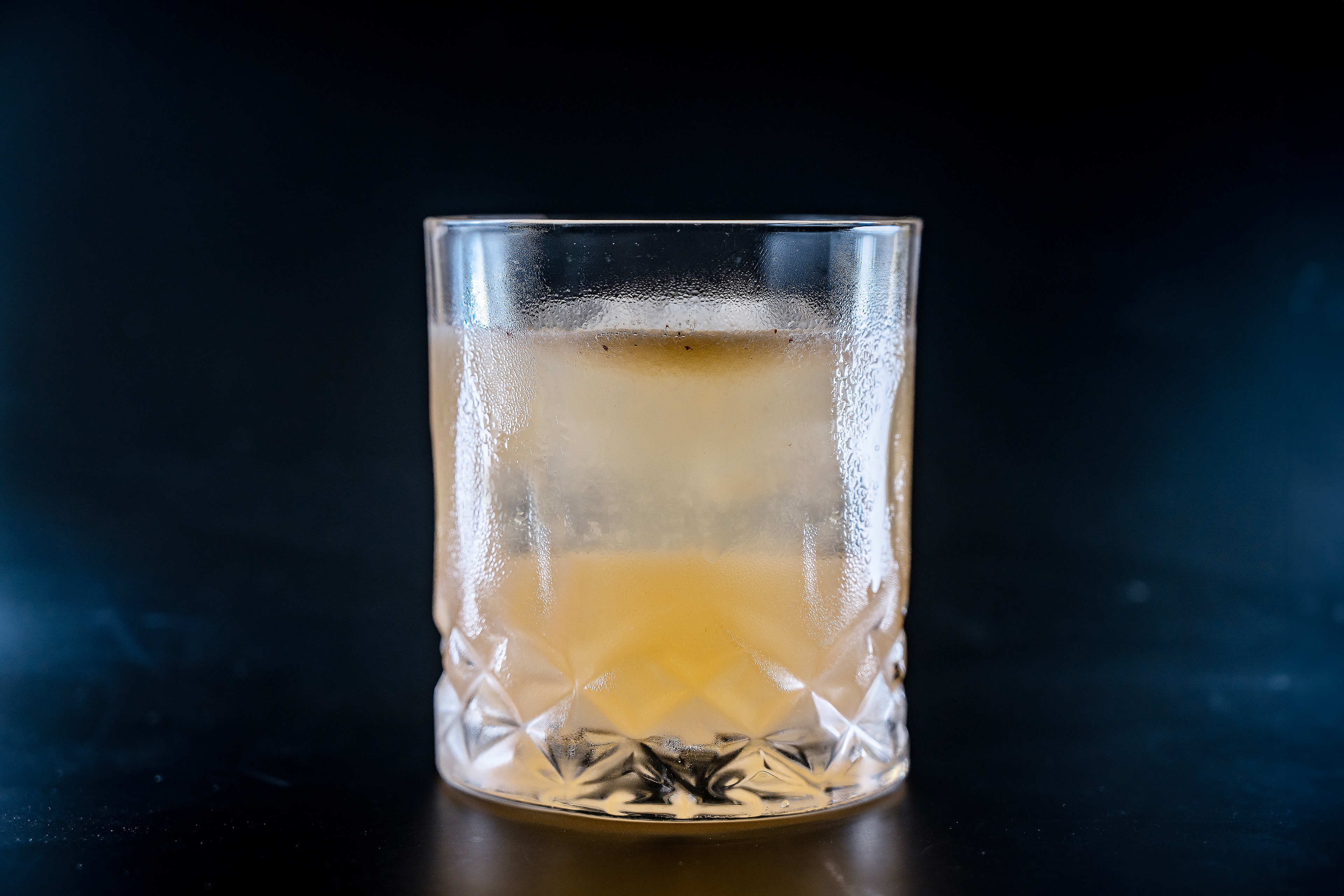 A vibrant Spicy Citrus Paloma cocktail served in a clear rocks glass over ice, set against a dramatic black background. The drink features a rich pink hue from fresh grapefruit and lime juices, subtly infused with chili flakes for a gentle heat. The minimalist presentation highlights the bold flavors of tequila, making this cocktail visually striking and perfect for those seeking a unique and refreshing tequila recipe.