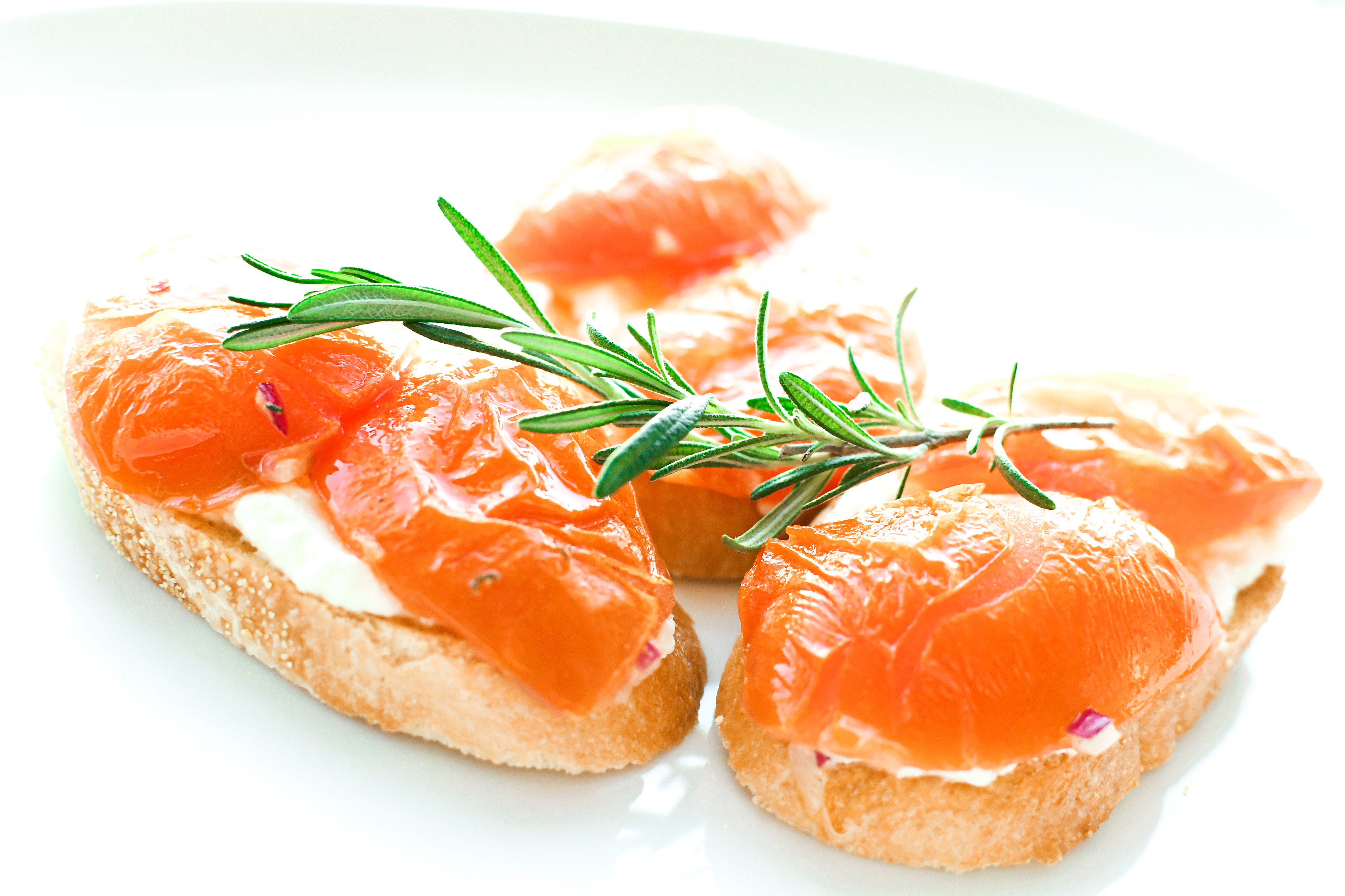 A white plate with roasted tomato bruschetta arranged on it, featuring slices of baguette topped with homemade ricotta cheese and roasted tomato quarters. The bruschetta is garnished with a sprinkle of freshly ground black pepper. The dish is set against a plain white background, highlighting the simplicity and freshness of the appetizer.