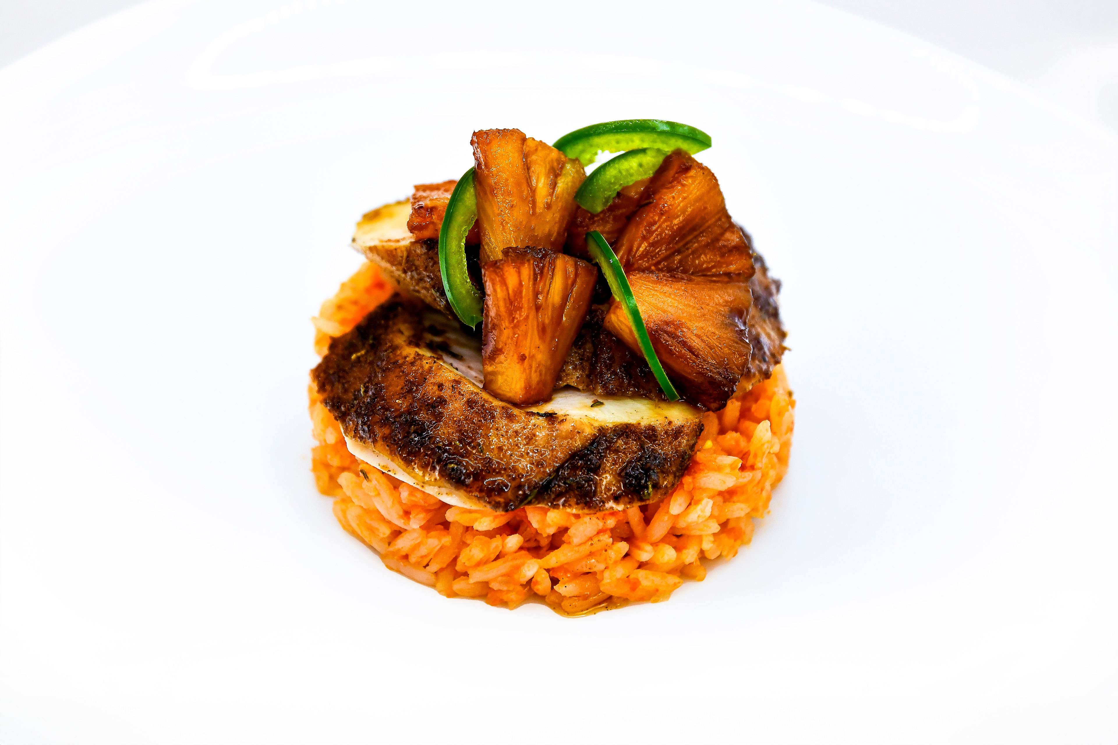 A beautifully plated dish of Jamaican jerk chicken served on a bed of vibrant orange rice. The chicken is topped with caramelized pineapple chunks and sliced jalapeños, adding a touch of color and texture to the presentation. The dish is set against a clean white background, emphasizing its rich and appetizing appearance.
