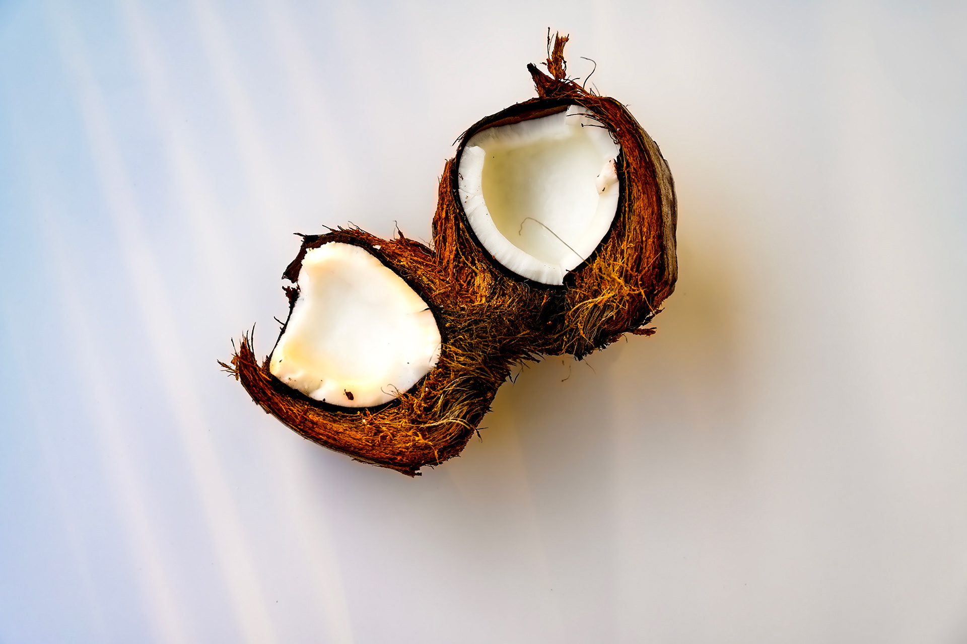 A raw coconut split open on a white background, with its fibrous brown husk still attached, highlighting the dramatic texture and natural beauty of the fruit.