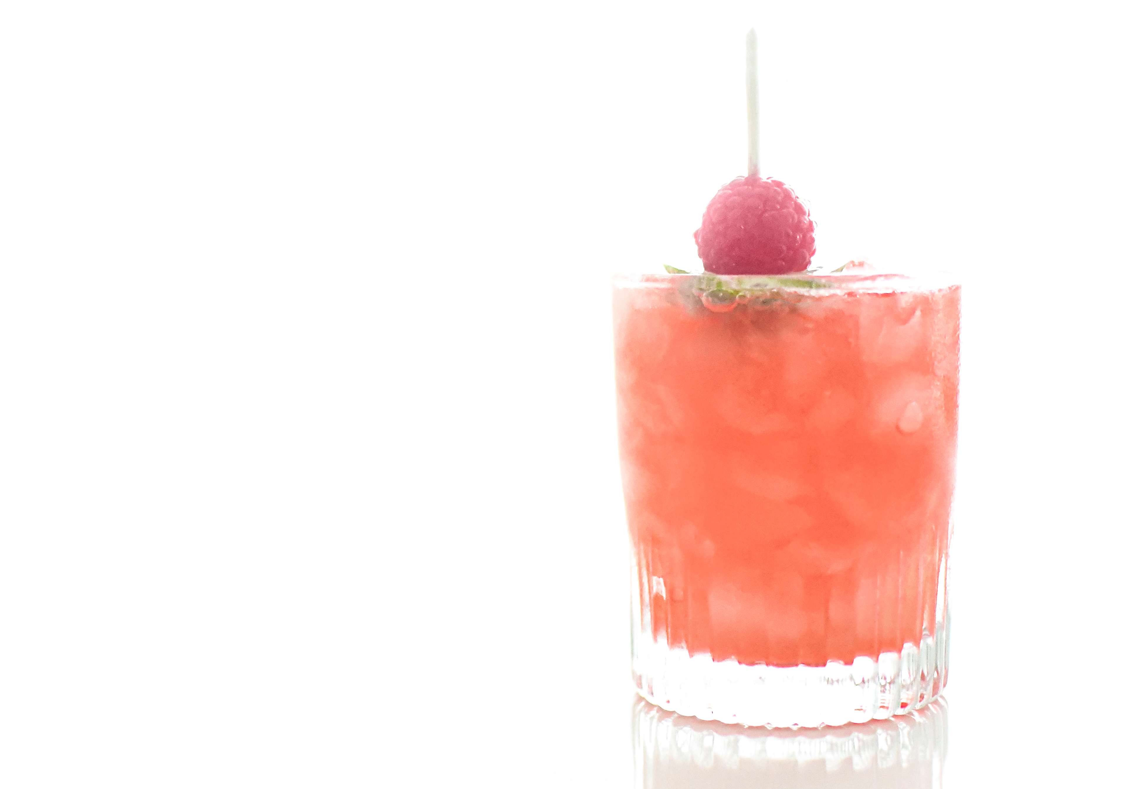 A dreamy, slightly distant view of a Knickerbocker cocktail on a white background. The rock glass is filled with crushed ice, and the cocktail is garnished with a lime wheel and a fresh raspberry. The image captures the entire glass, showcasing the vibrant, refreshing drink with a soft, elegant focus, emphasizing the cocktail’s visual appeal and the chilled, inviting presentation.