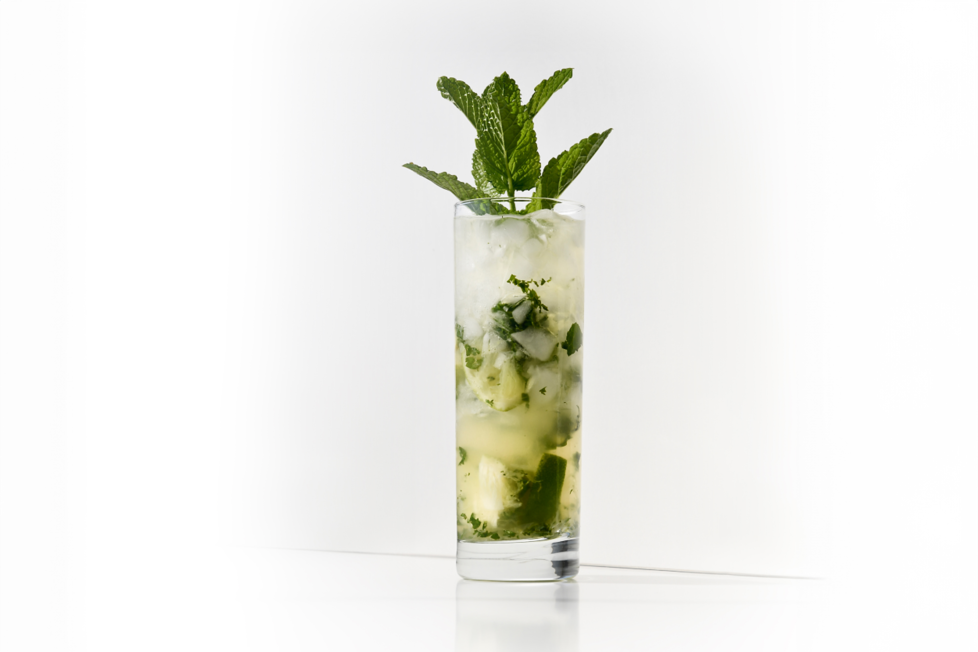 A classic Mojito cocktail on a white background. The highball glass is filled with lime wedges, mint leaves, and crystalline white sugar, showcasing the fresh and vibrant ingredients. The cocktail is prepared with Ron Botran Reserva Blanca and topped with mineral water, garnished with a mint sprig. The image highlights the refreshing and aromatic qualities of the Mojito, emphasizing the classic preparation steps and ingredients.