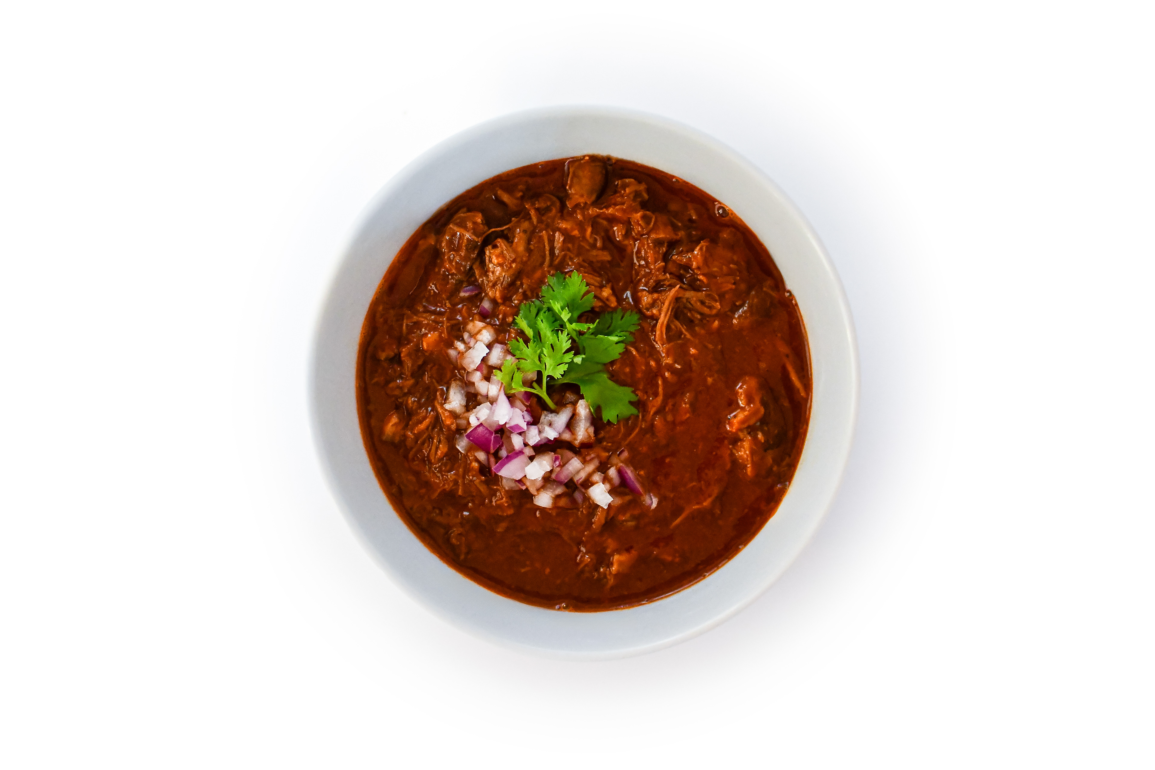 A white bowl filled with tender, shredded beef birria in a rich, red broth. The birria is garnished with fresh cilantro, creating a vibrant contrast against the neutral white background, highlighting the dish's smoky and savory flavors.