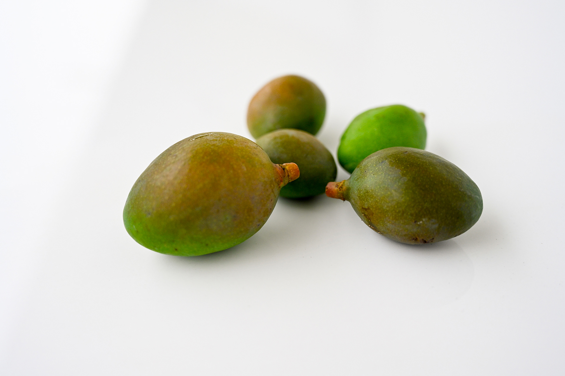 A spontaneous arrangement of five young, unripe mangoes scattered randomly on a neutral-toned surface, highlighting natural imperfection and freedom.