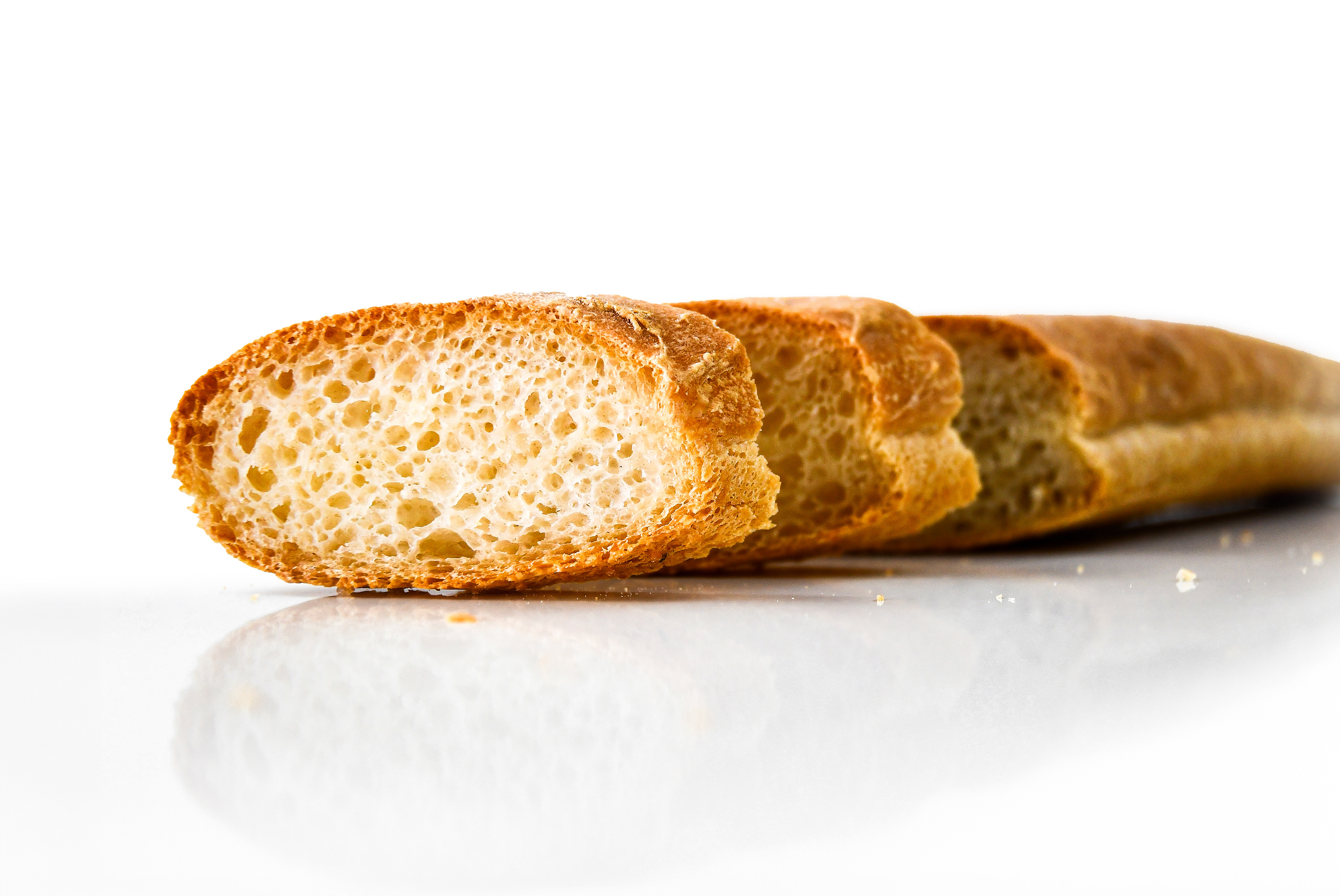 A baguette on a white, shiny surface. The baguette is whole with two pieces cut from the front, showcasing its crispy crust and interior. The baguette's reflection is visible on the surface.