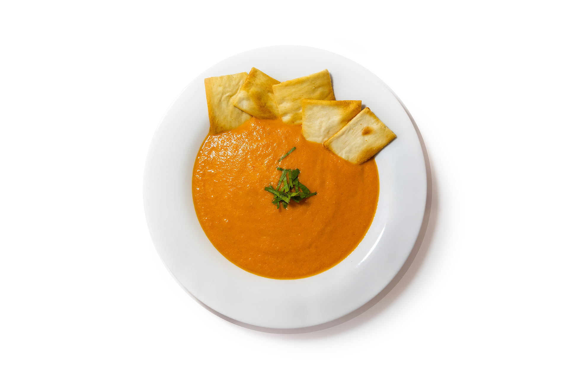 A vibrant red-orange tomato soup served in a white bowl on a white background, garnished with fresh herbs and accompanied by crisp crackers on the side.