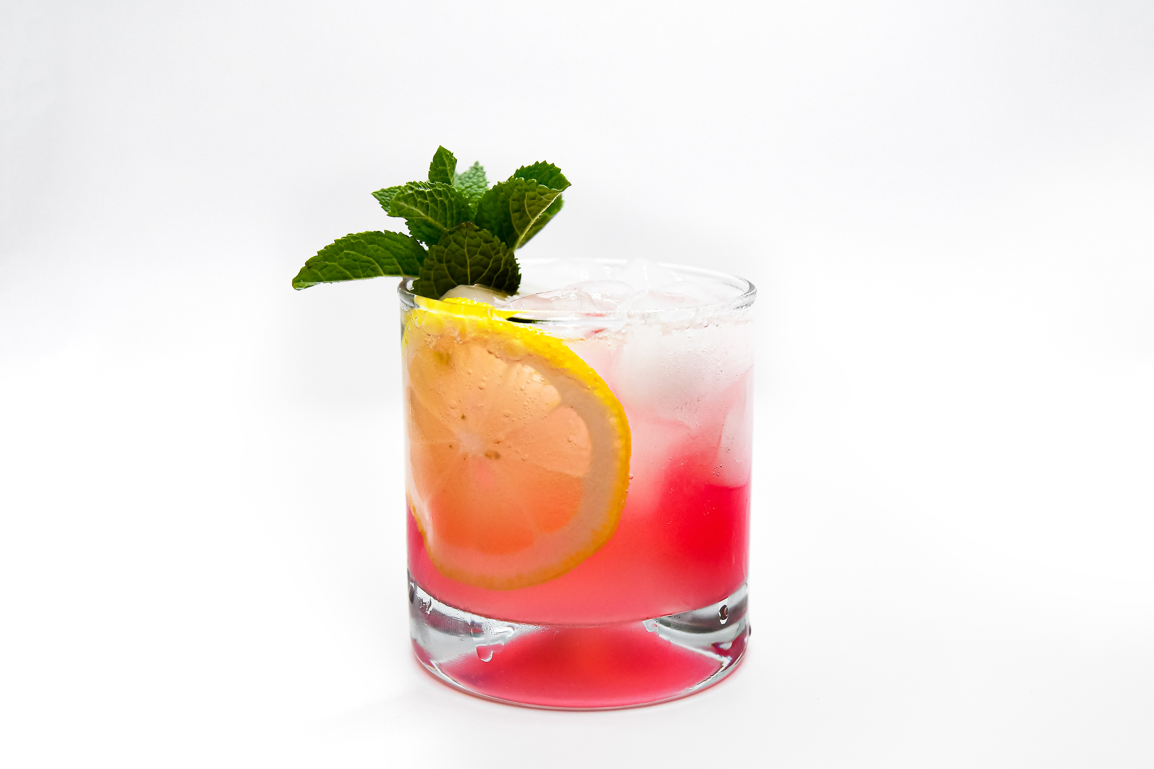 A Gin Daisy cocktail served in a sleek rock glass, showcasing the refreshing blend of gin, lemon juice, homemade grenadine, and topped with a splash of soda water. The image highlights the elegant presentation of the drink, focusing on the vibrant red hue at the bottom transitioning to clear at the top. Inside the glass, a lemon slice and a sprig of mint are elegantly displayed, while the ice cubes add to the cocktail’s chilled appearance. The white background accentuates the vibrant colors and simple, refreshing appeal of the drink.