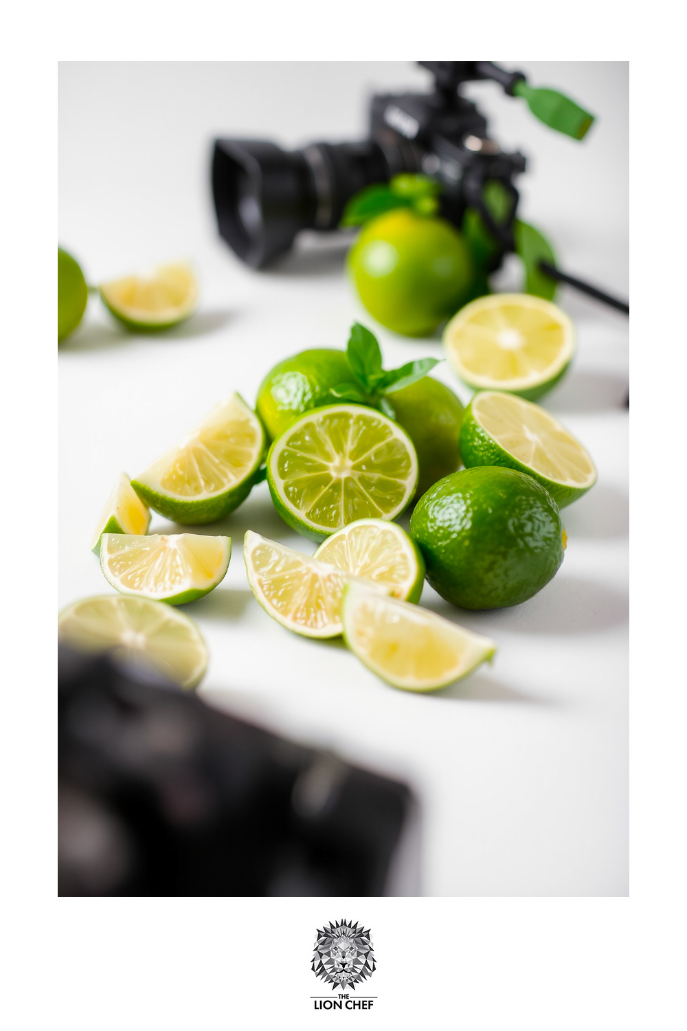 A behind-the-scenes photo shoot setup featuring a camera alongside fresh limes, sprigs of mint, and a small pile of sugar, all arranged on a clean white background. The vibrant green of the limes and mint contrasts with the white surface, highlighting the fresh ingredients used for cocktail preparation
